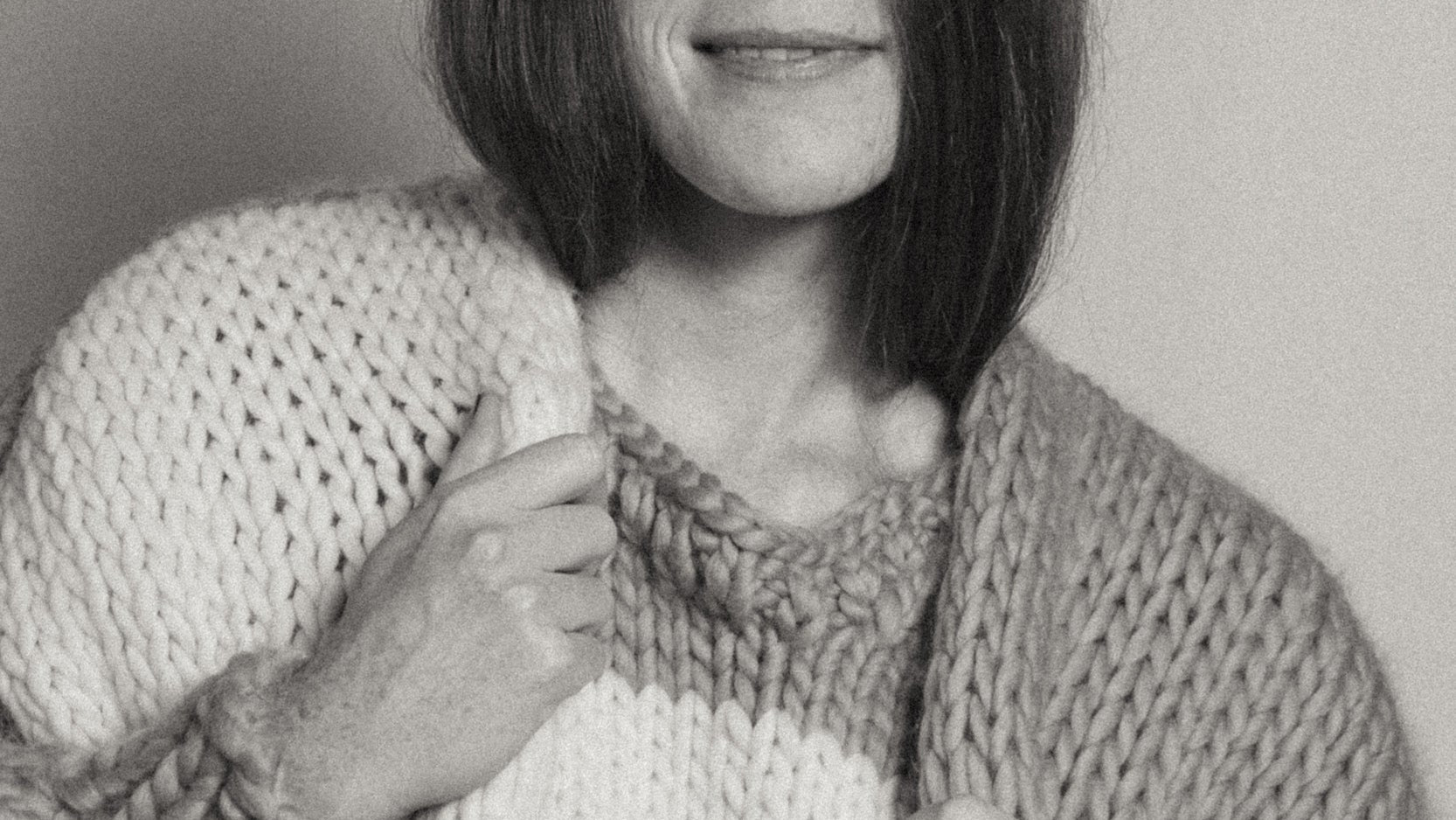 A close-up black and white photo of a person wearing a chunky knit sweater and cardigan, holding the collar area with one hand. The image is monochrome, showing shades of gray, white, and black.