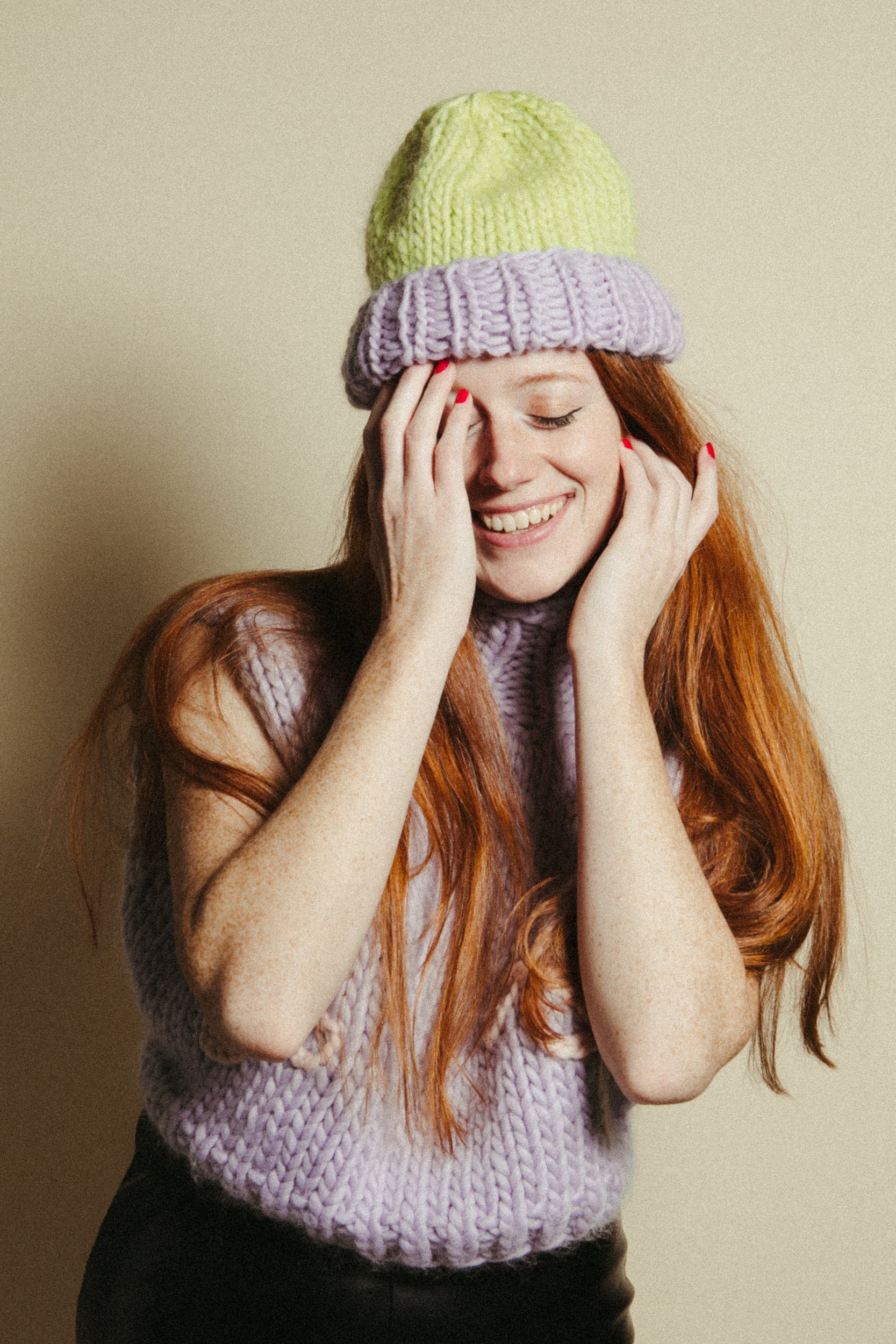 Woman wearing a green knit beanie and purple knitted sleeveless sweater against a beige background