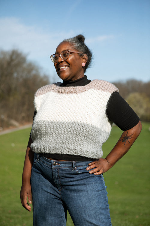 Person wearing a three-tone sweater and jeans standing outdoors with trees and sky in the background