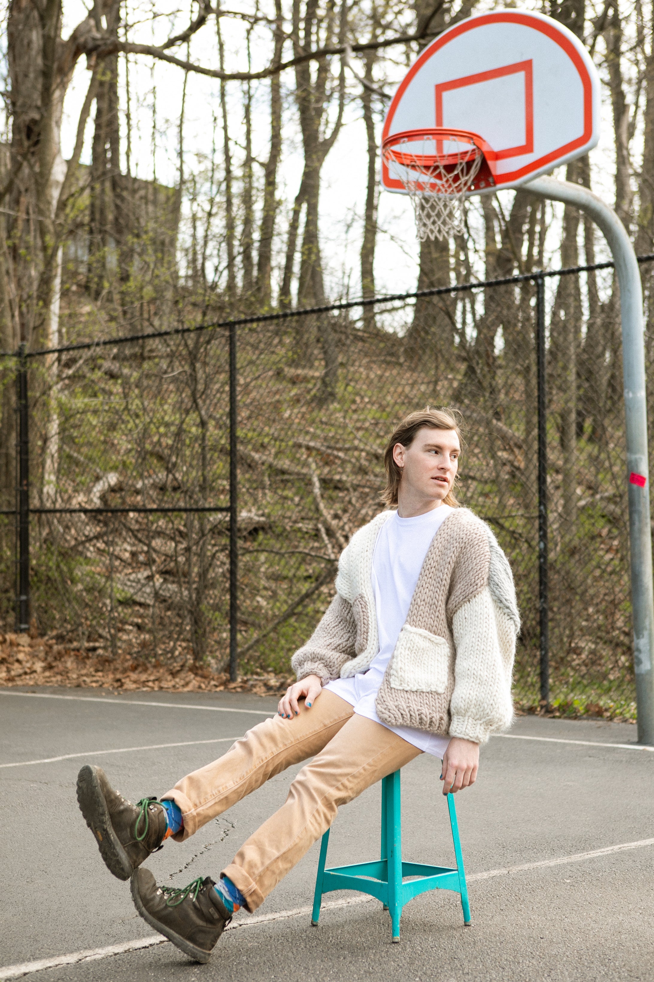 Person wearing a cardigan sitting on a blue stool in front of a basketball hoop on an outdoor court.