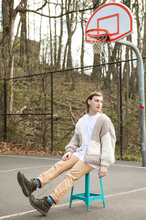 Person wearing a cardigan sitting on a blue stool in front of a basketball hoop on an outdoor court.