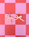 A pair of chunky knit bow earrings in a pale peach-pink color with gold-tone fishhook ear wires, displayed on a pink and red checkered background.