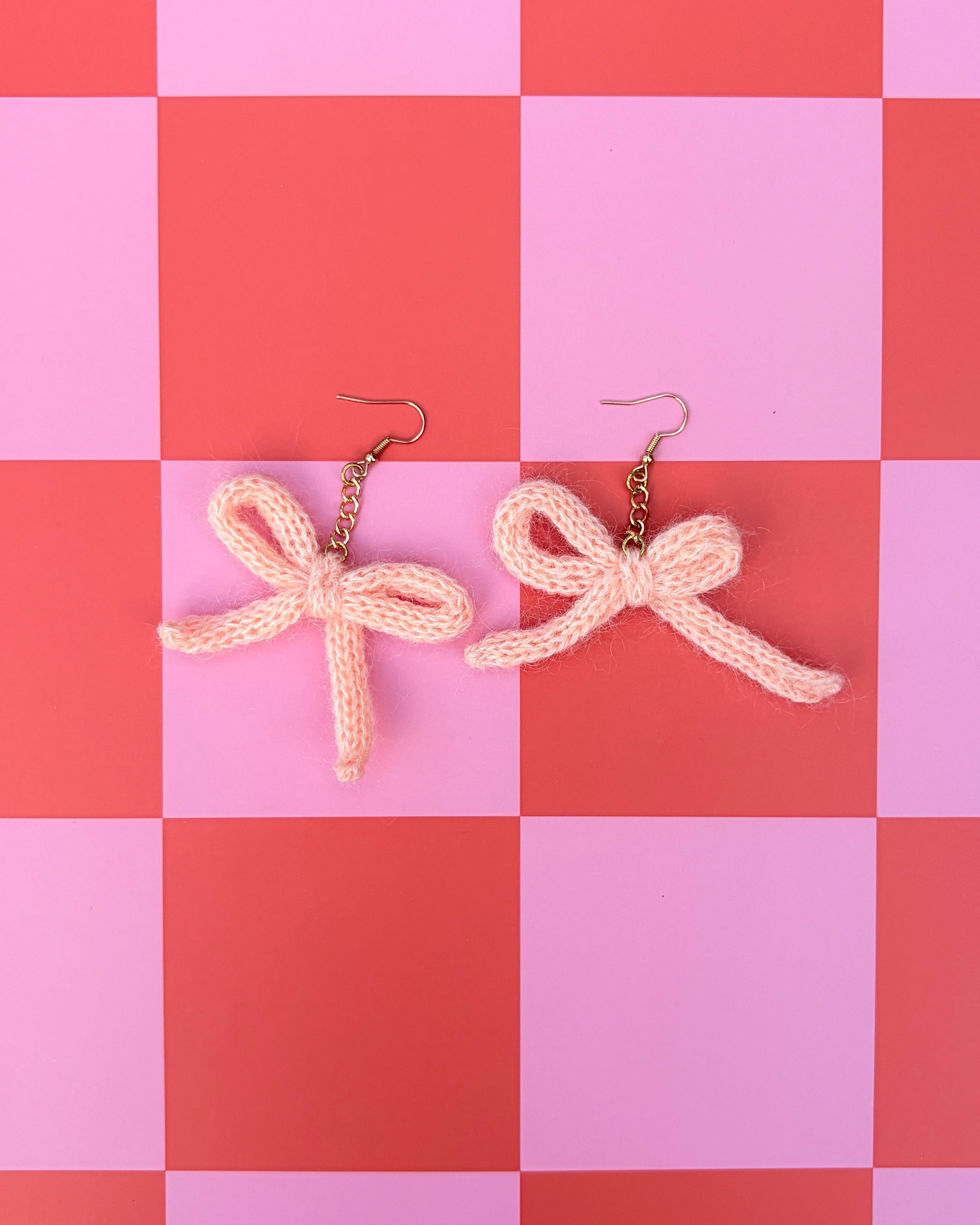 A pair of chunky knit bow earrings in a pale peach-pink color with gold-tone fishhook ear wires, displayed on a pink and red checkered background.