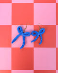 A pair of bright blue knitted bow earrings with gold fishhook wires, displayed on a pink and orange checkered background.
