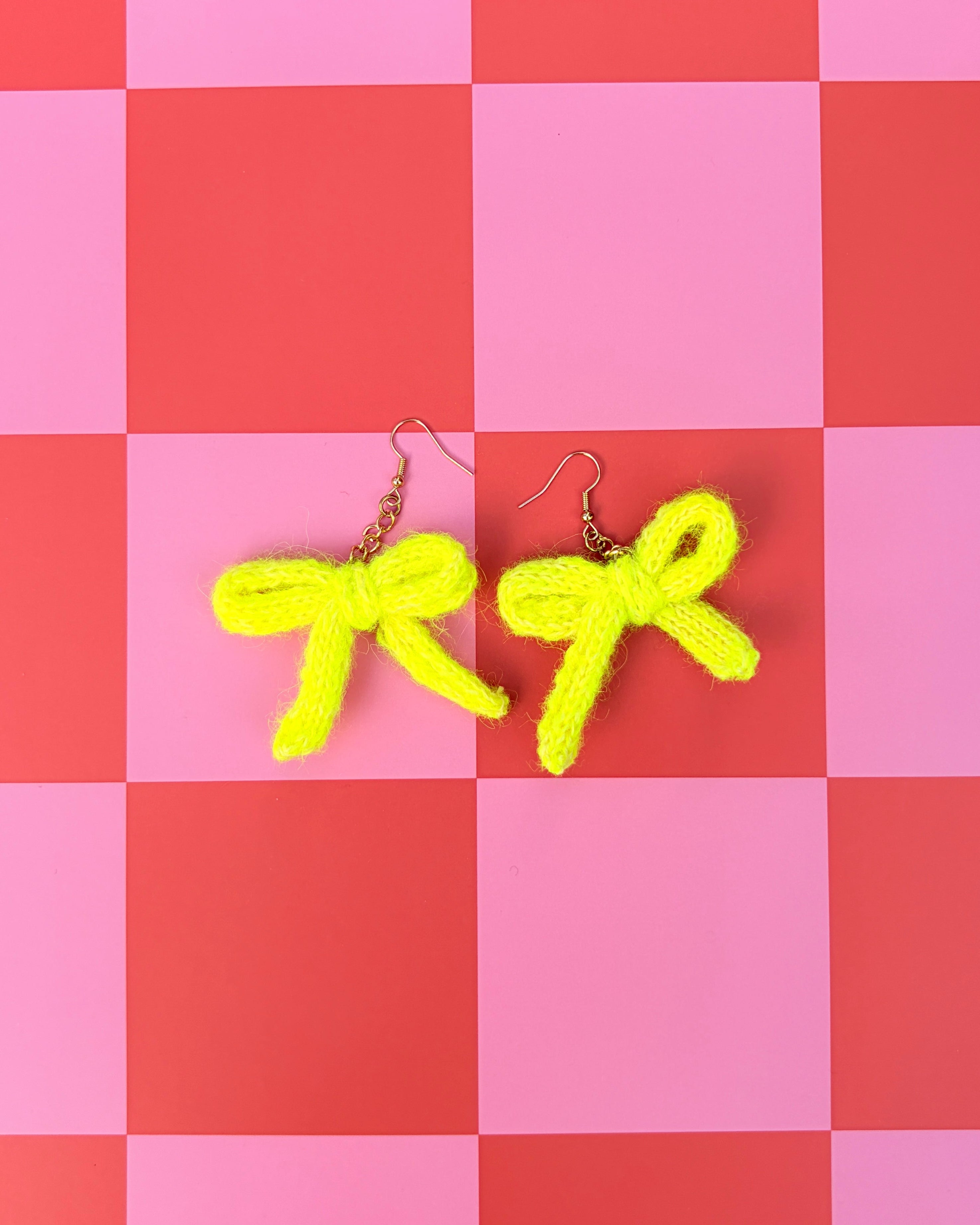 A pair of bright yellow fuzzy bow-shaped earrings with gold fishhook wires, displayed on a pink and red checkered background.
